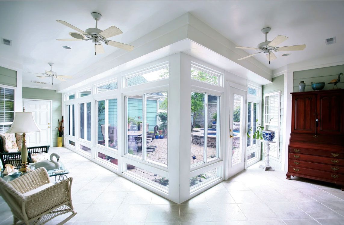 A brightly lit sunroom with large windows, white walls, ceiling fans, tiled floors, a wicker chair, and a wooden hutch.