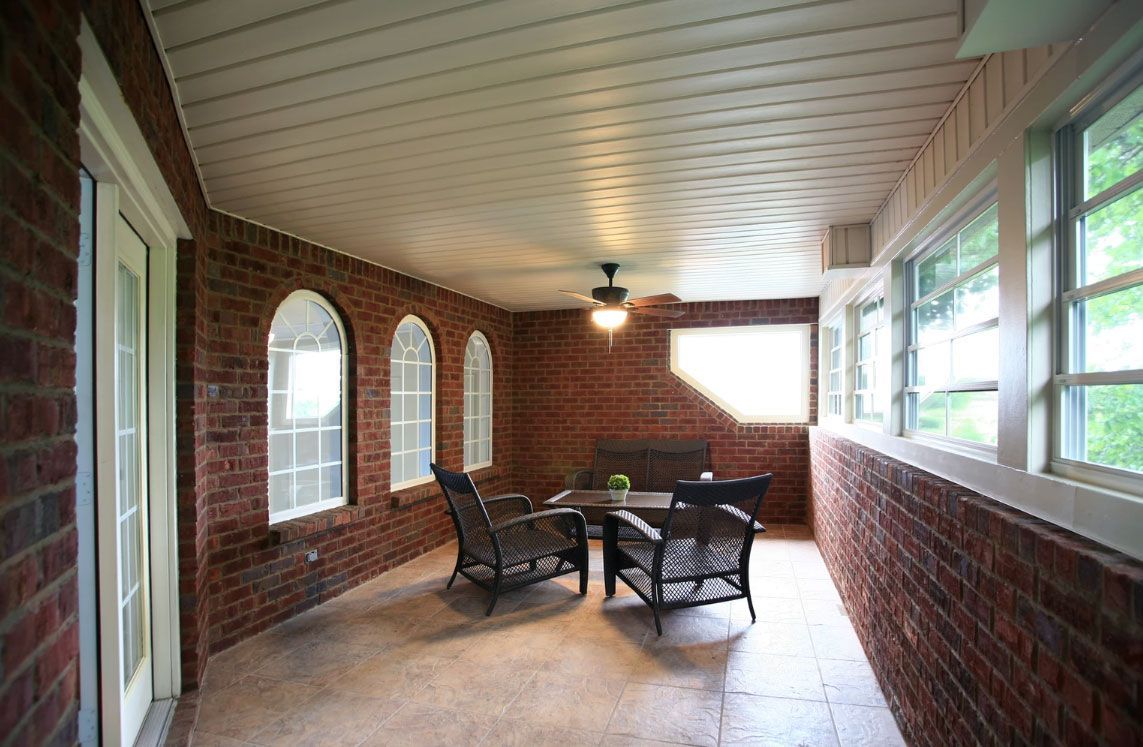 A sunroom with brick walls, tiled floors, a ceiling fan, and two armchairs facing a small table.