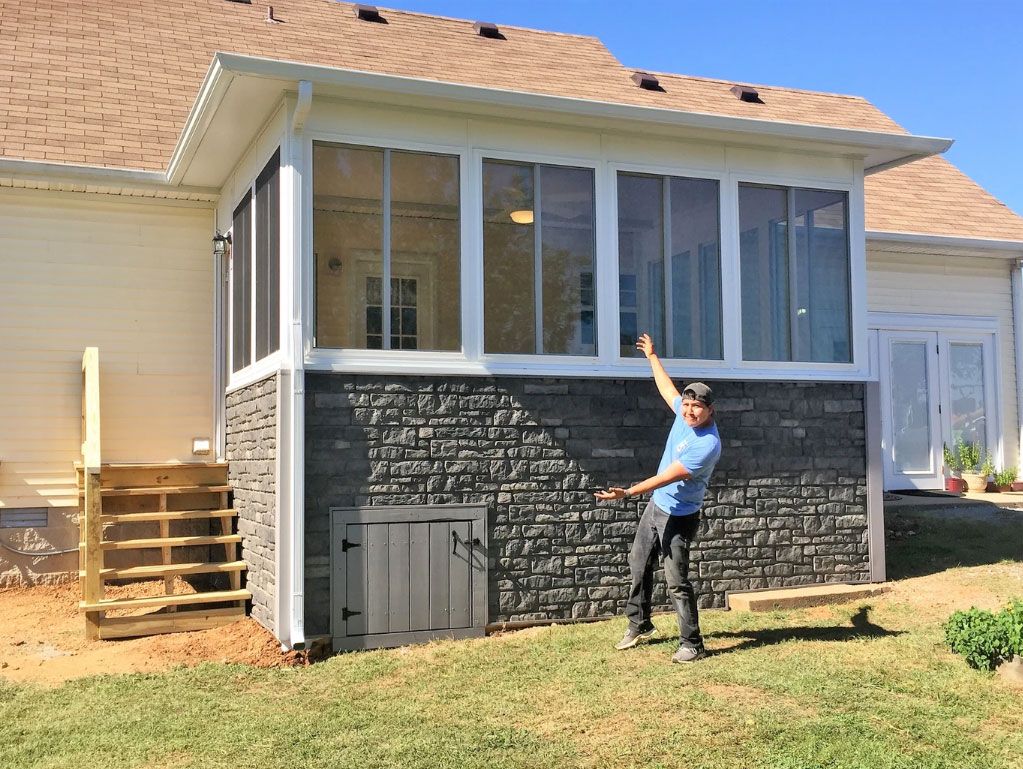 A person in a blue shirt points to a new sunroom extension with stone-textured skirting, attached to a light-colored house.
