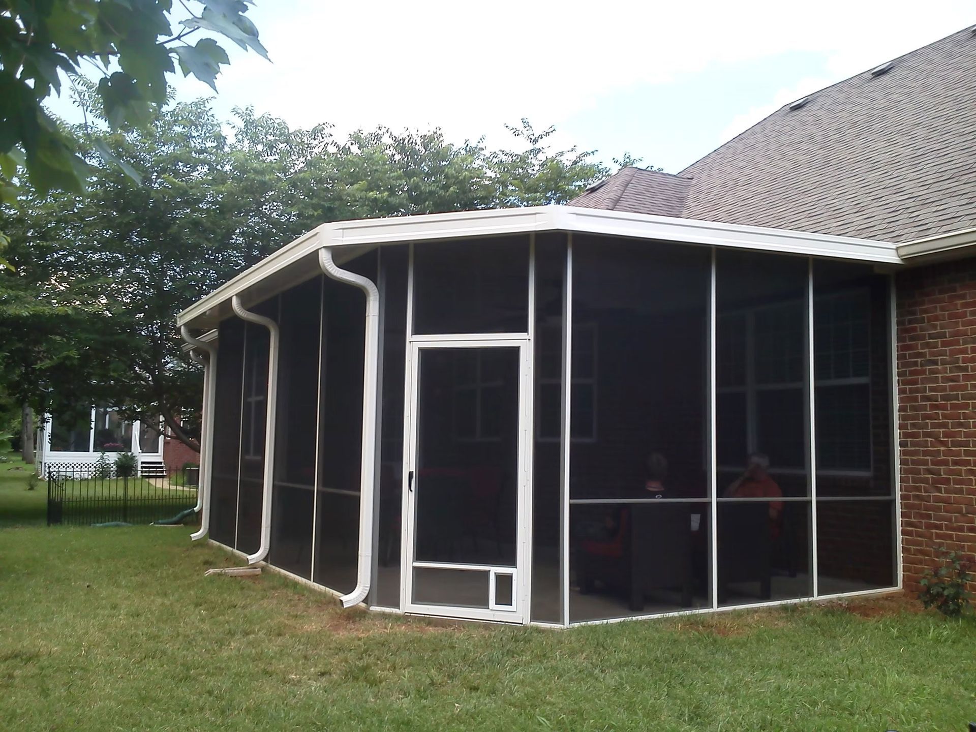 A white-framed screened-in porch attached to the brick exterior of a house, set in a grassy backyard with trees.