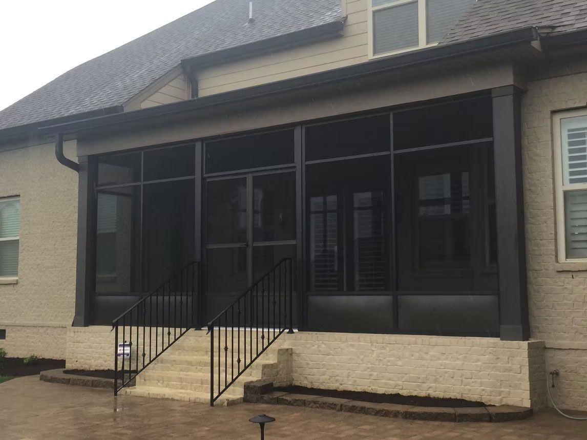A beige brick house features a black-framed, covered screened-in patio with stairs leading down to a stone patio.