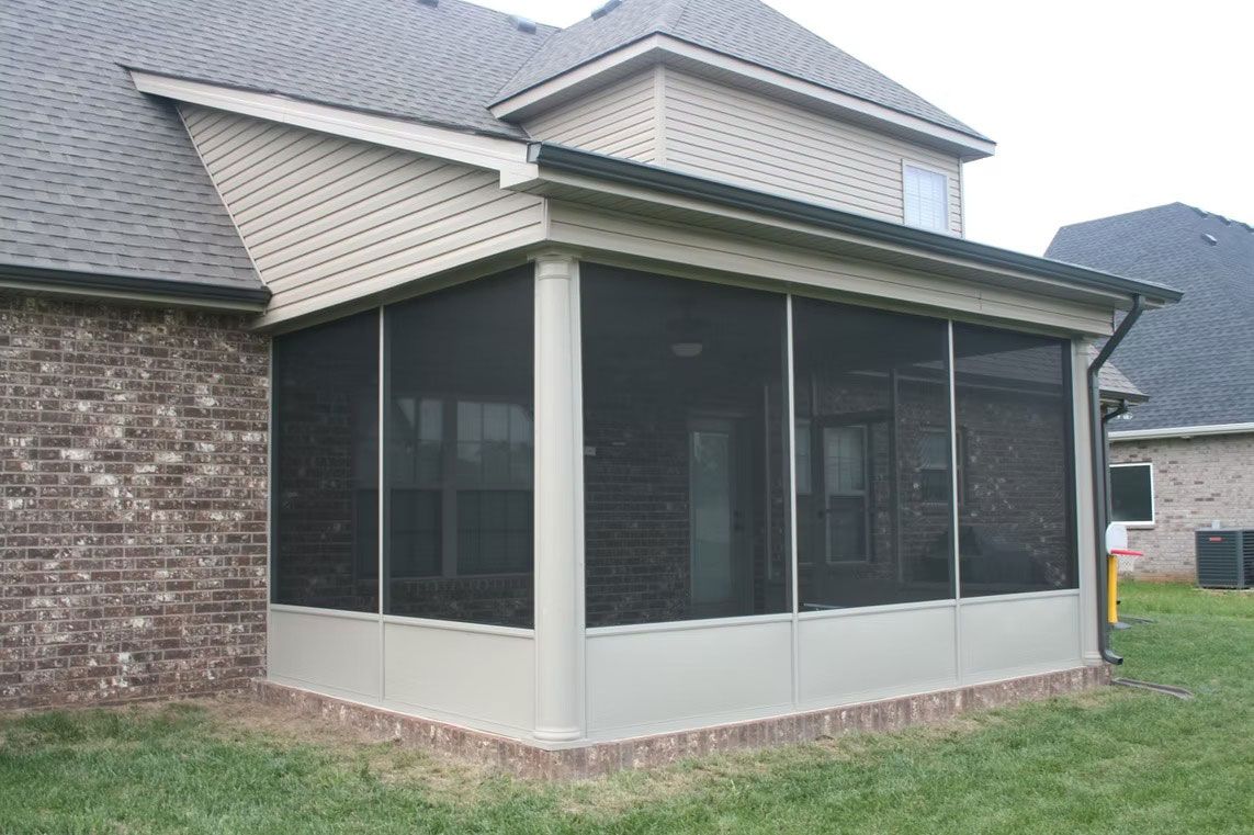 A tan screened-in porch addition attached to a brick house with a gray shingled roof in a backyard setting.