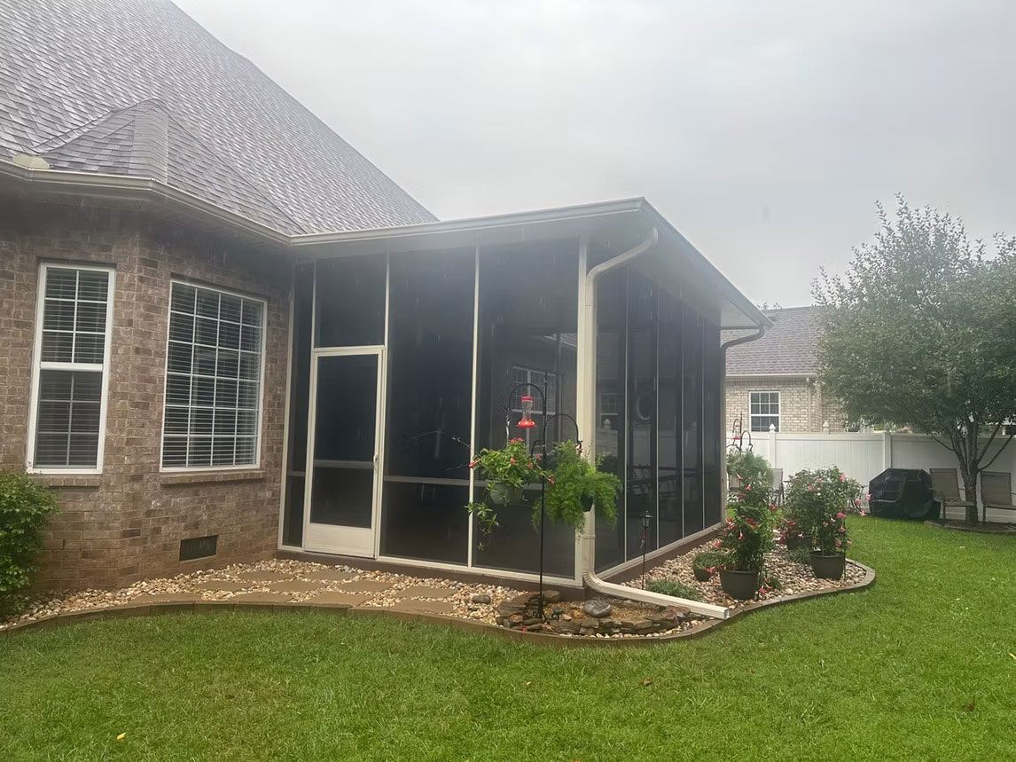 A brick house exterior with a screened-in porch, adjacent patio area with small potted plants, and a grassy lawn.
