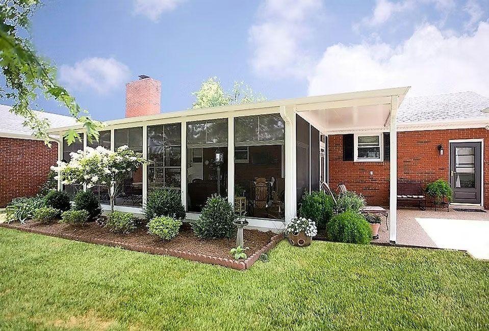 A red brick home featuring a white-framed screened-in sunroom with a patio and manicured landscaping in the backyard.