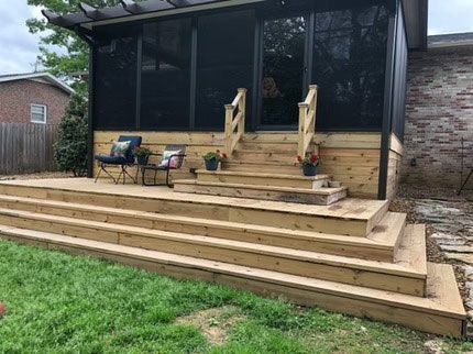 A wooden deck with wide steps leading to a screened-in porch, featuring two chairs and potted plants on a sunny day.