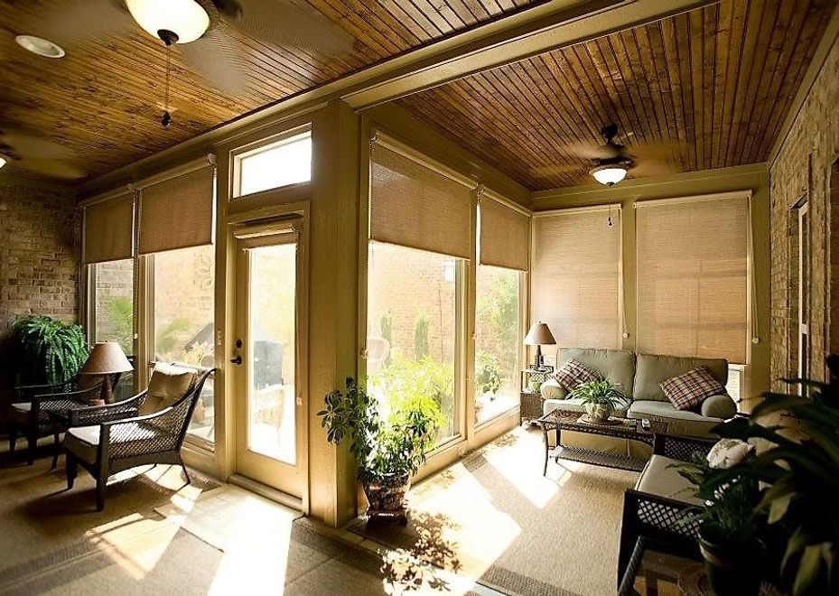A sunroom with a wood-paneled ceiling, large windows with roller shades, rattan furniture, and potted plants.
