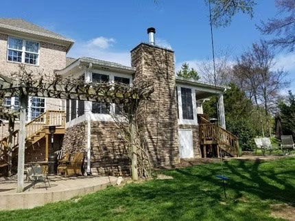 A multi-story brick and stone house with a stone chimney, a wooden deck, a pergola, and a grassy lawn on a sunny day.
