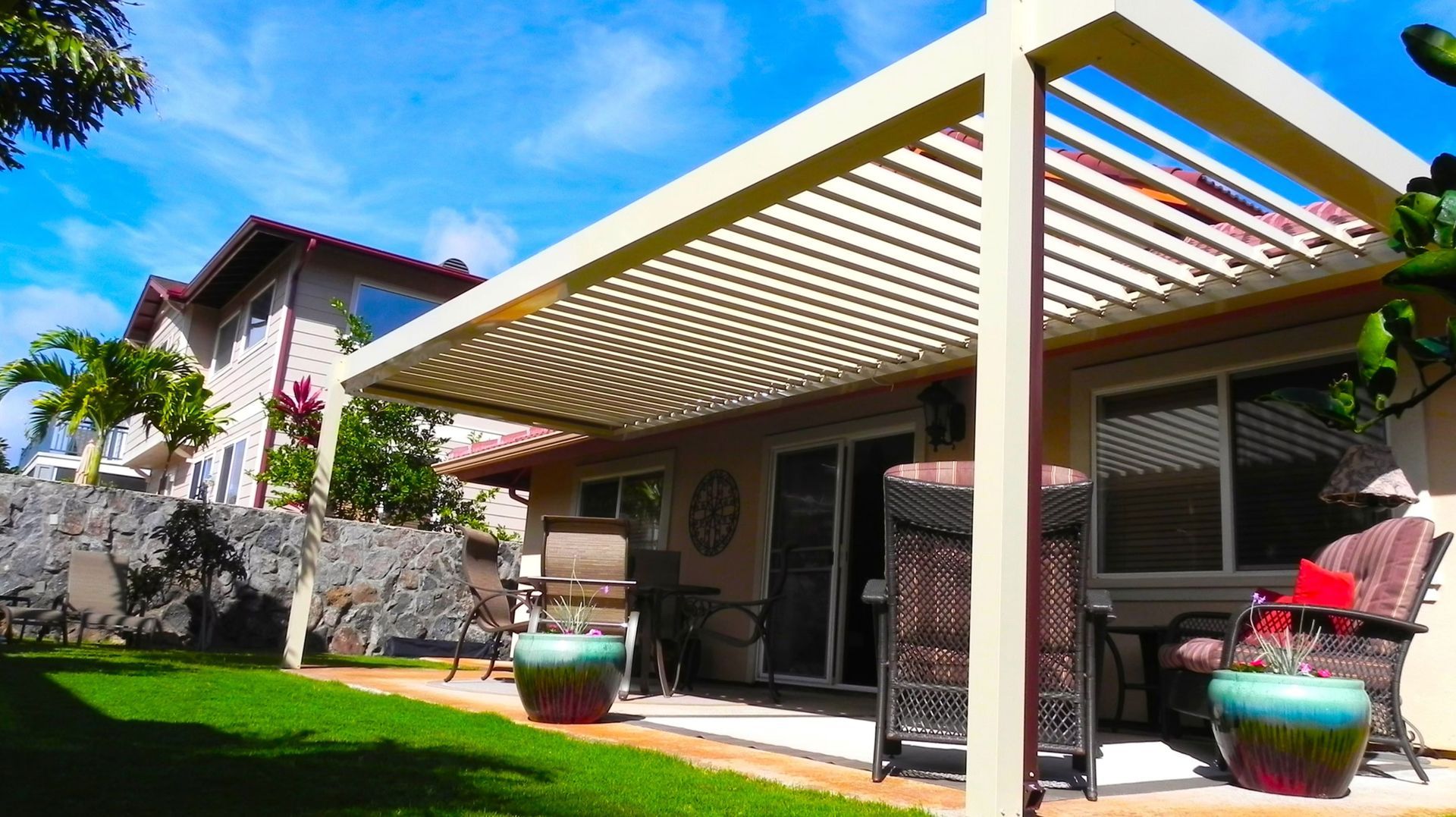A patio with a modern louvered pergola, outdoor furniture, and colorful planters set against a house and lush backyard.