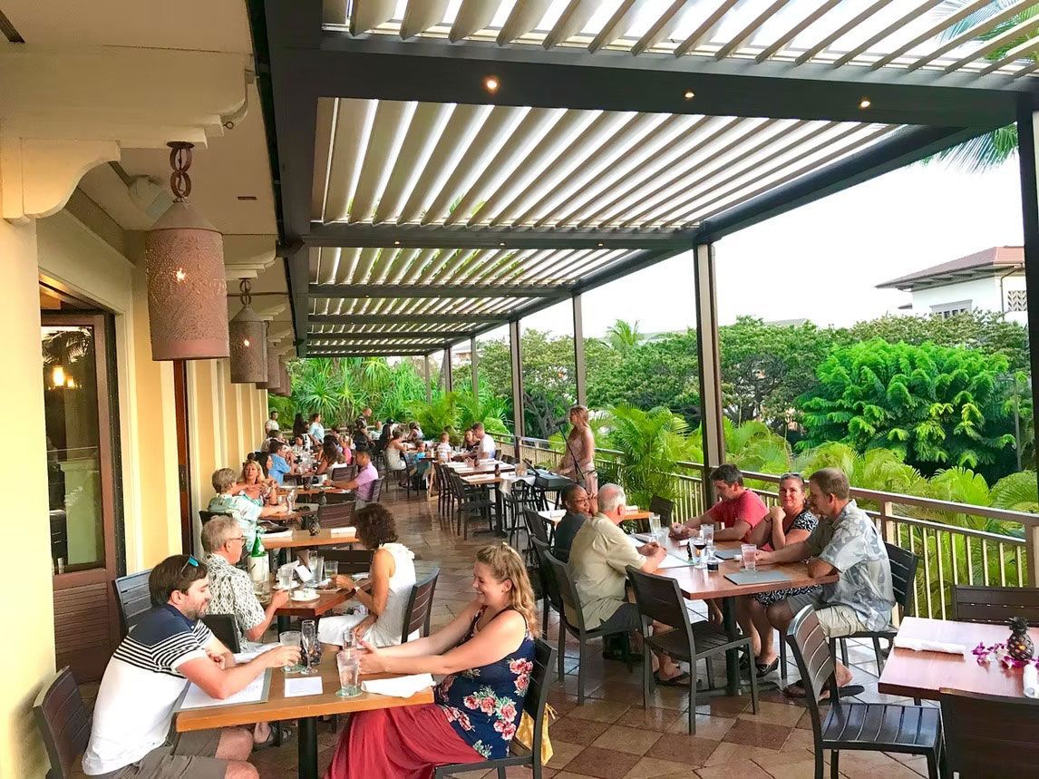 People dine at tables under a shaded, open-air patio with a slatted roof overlooking lush green landscaping.