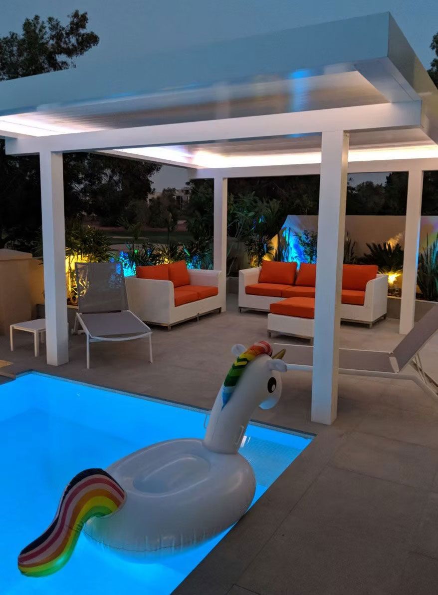 A poolside lounge area at dusk featuring white patio furniture with orange cushions under a modern, illuminated pergola.