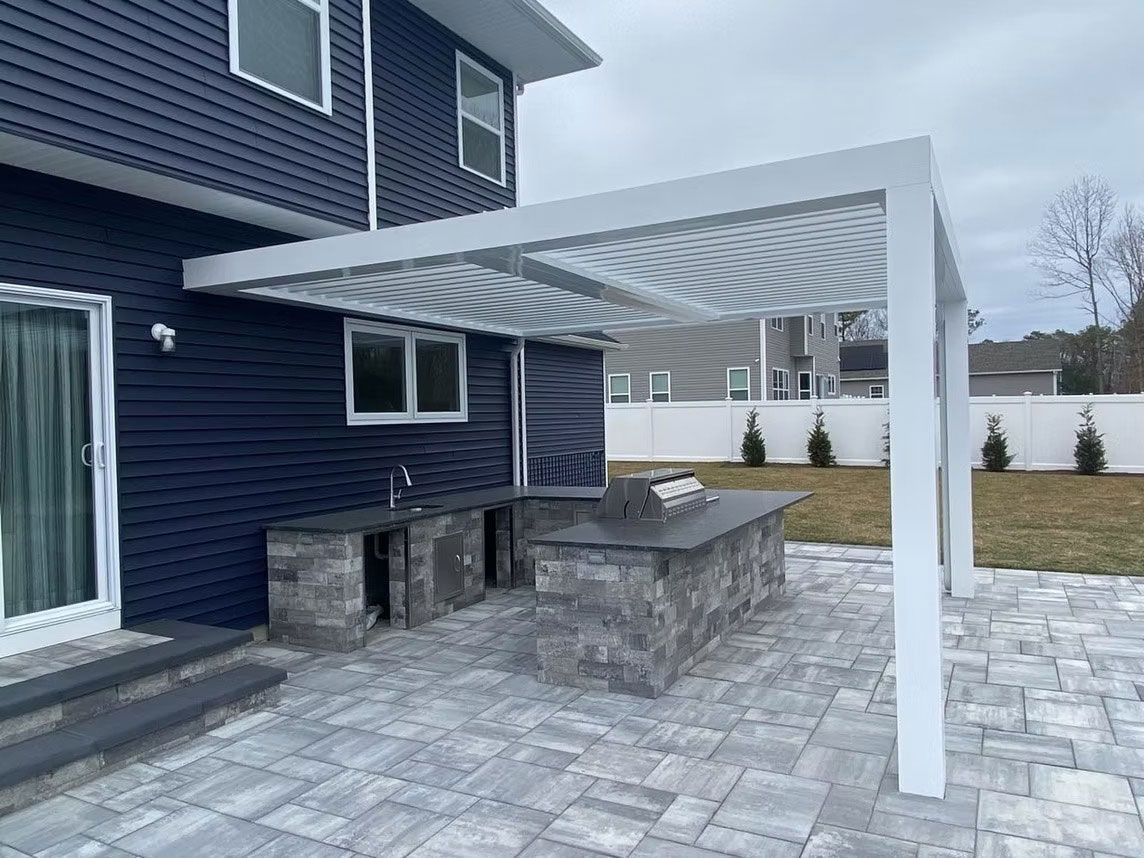 Outdoor kitchen with stone counters and a white louvered pergola on a gray paver patio against a dark blue house.