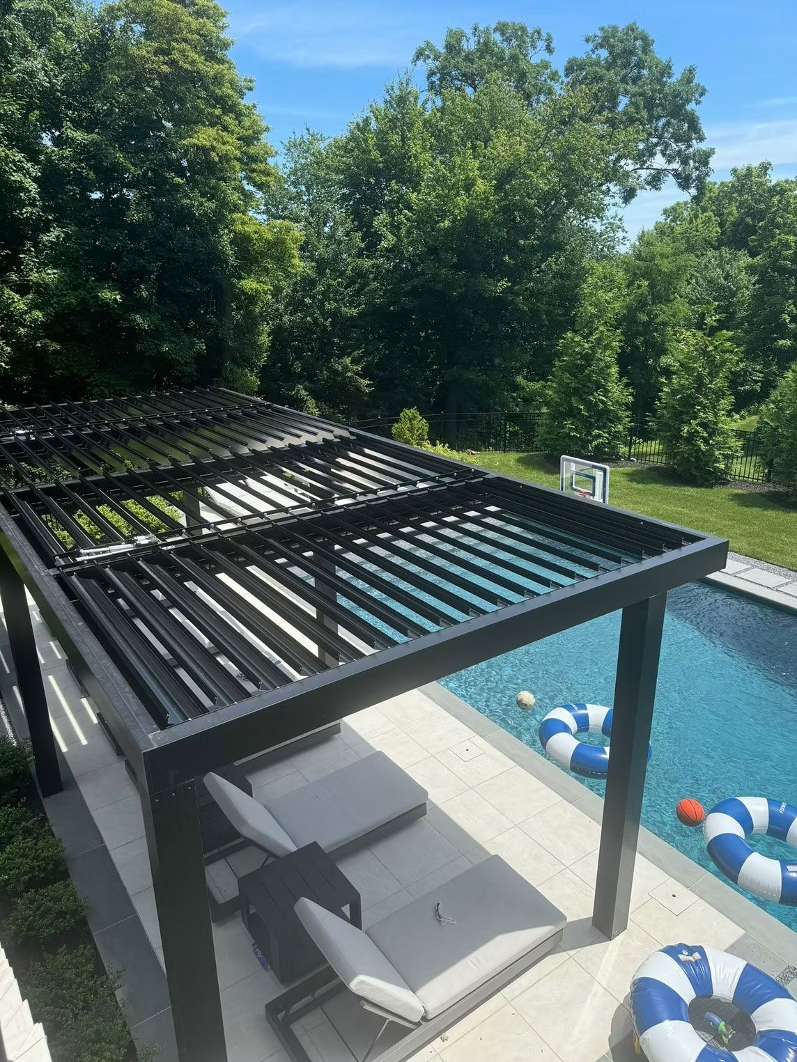 A modern black pergola shades two lounge chairs on a light-colored pool deck beside a pool with blue and white inflatables.