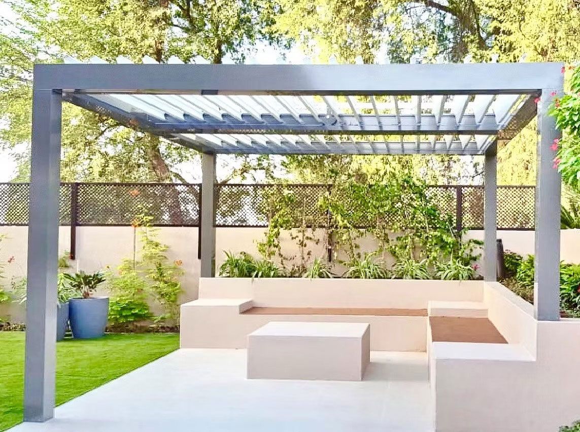 A modern grey pergola stands over a minimalist light-colored stone seating area in a sunny backyard garden.