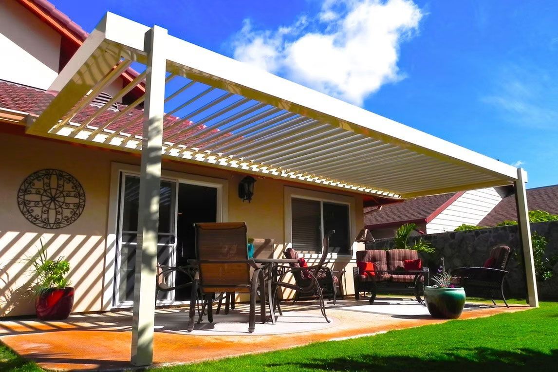 A patio featuring a white louvered pergola covering a dining table and chairs on a sunny day next to a house.