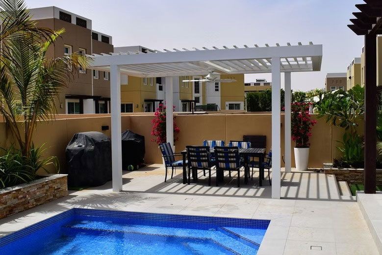 A patio with a white pergola, dining table, chairs, and two grills beside a blue swimming pool in a residential backyard.