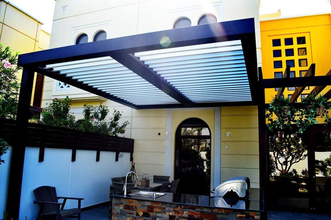 A patio featuring a dark metal pergola with a louvered roof over an outdoor kitchen, set against a two-story home.