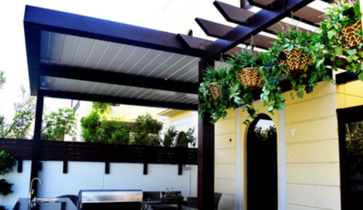 A patio with a dark wood pergola, a white paneled ceiling, a built-in outdoor kitchen, and hanging potted plants.