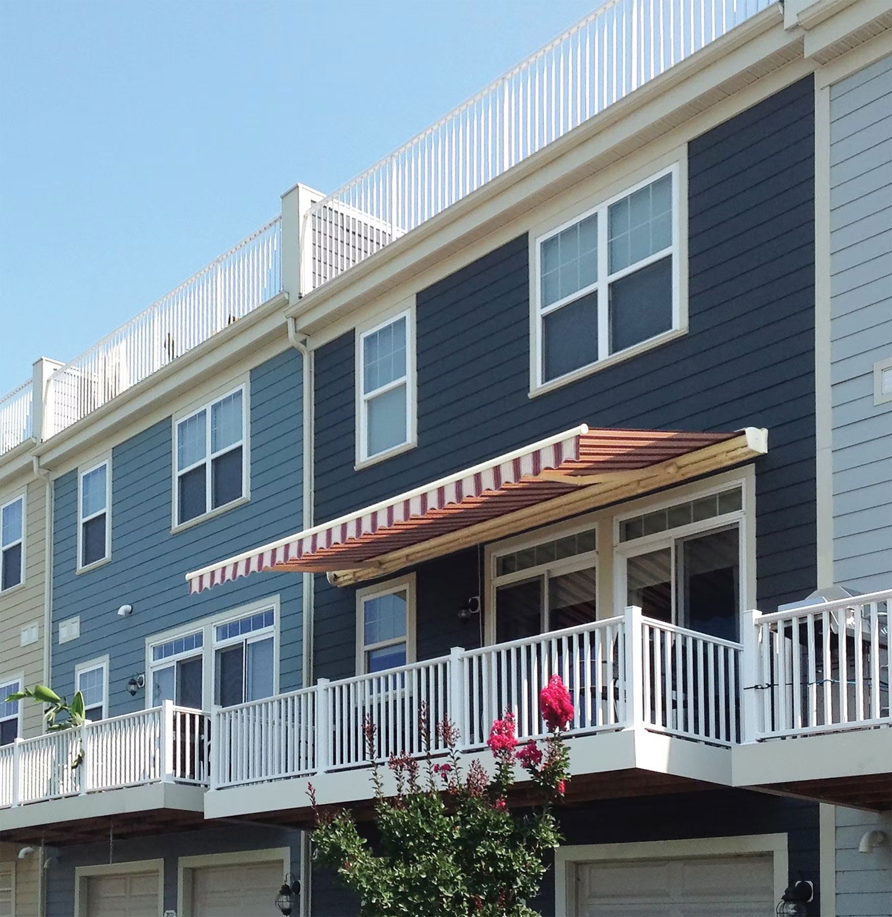 Dark blue, multi-story townhouses with white railings, balconies, and a striped retractable awning against a blue sky.