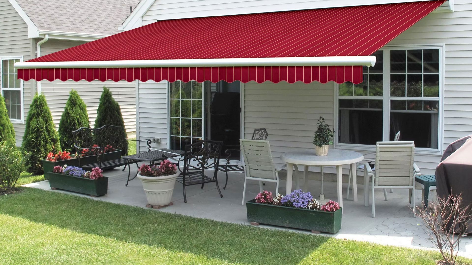 A red retractable awning extends over a concrete patio with a dining set and potted flowers against a white house.