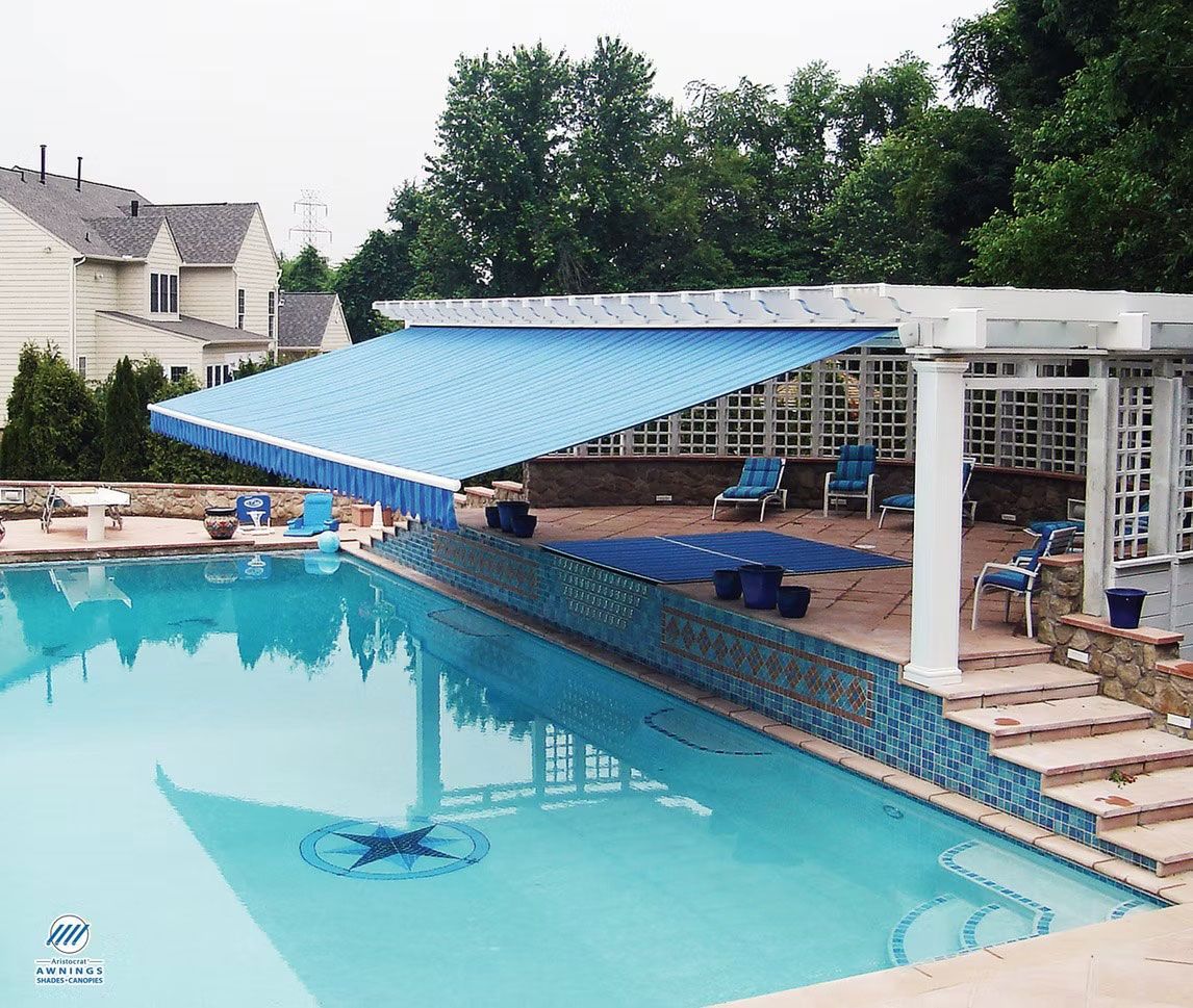Blue retractable awning extended over a backyard swimming pool patio with white columns and lounge chairs.