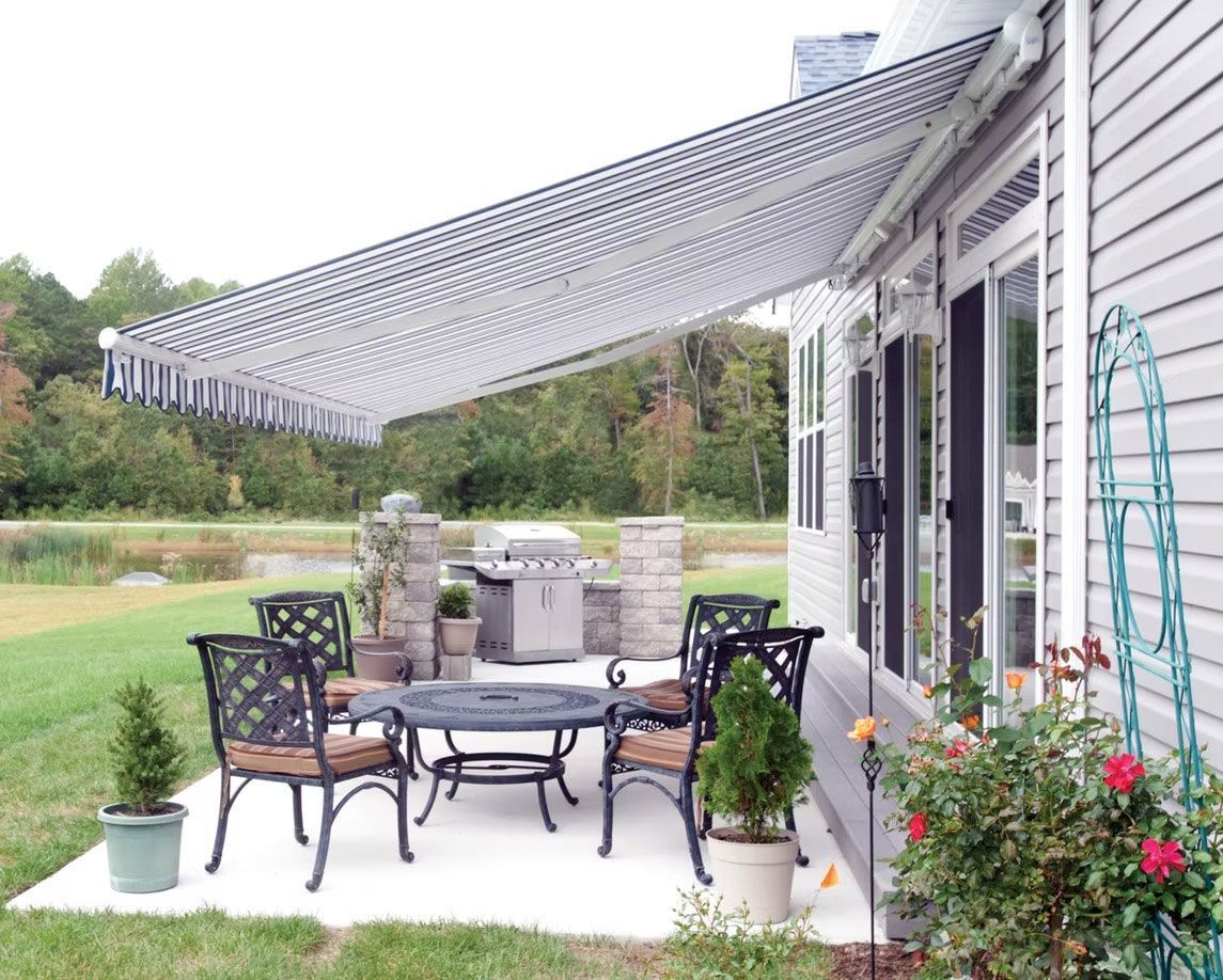 Patio with a black-and-white striped retractable awning, outdoor table and chairs, and a grill set on a grassy lawn.