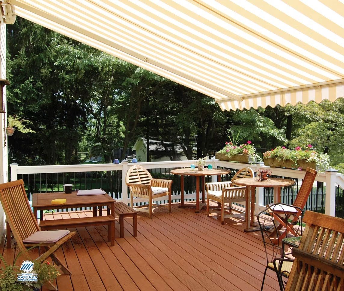 A wooden deck with patio furniture, including tables and chairs, under a striped retractable awning overlooking trees.