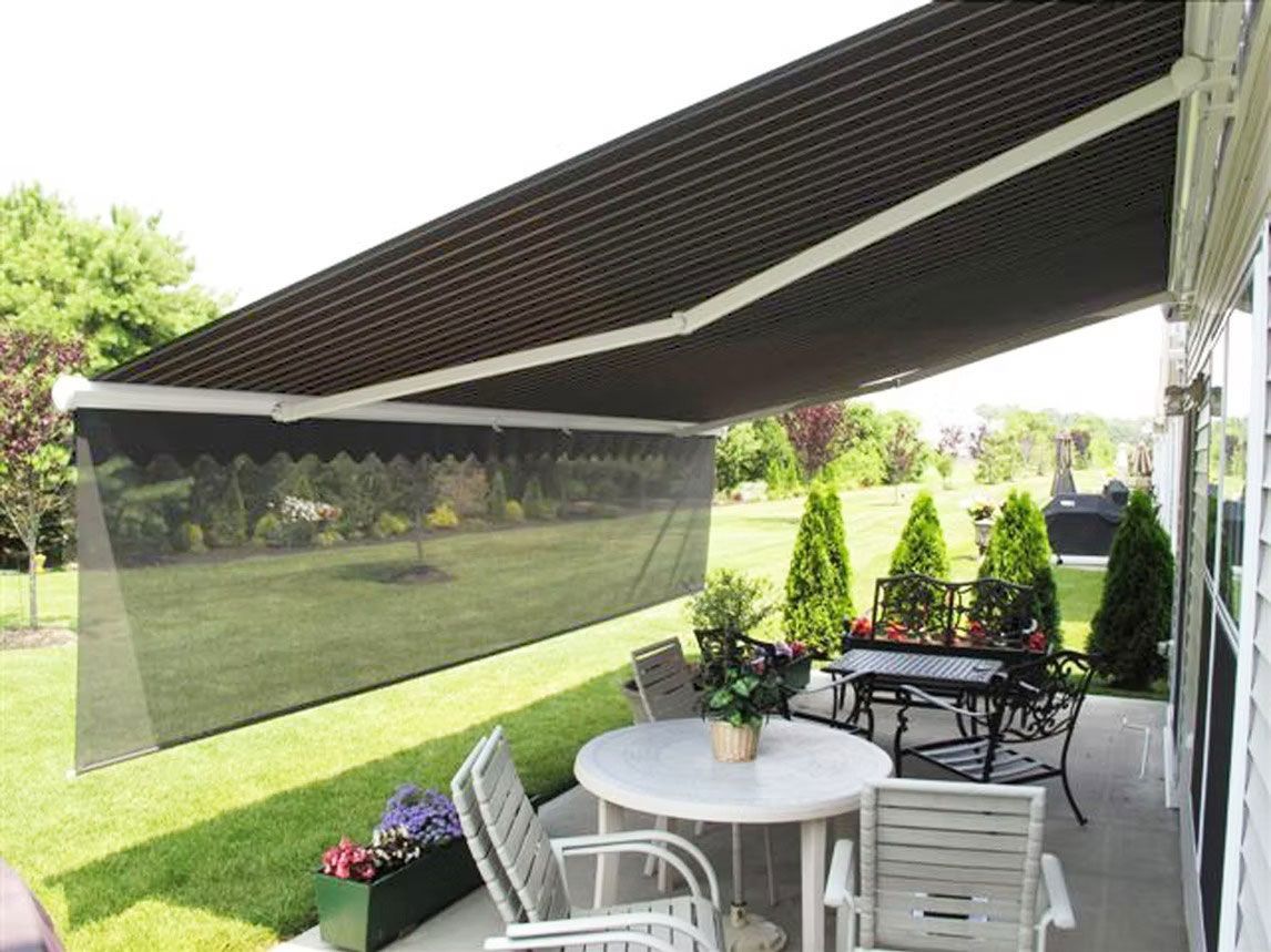 A retractable dark brown patio awning with a drop-down mesh shade over a table and chair set on a backyard patio.