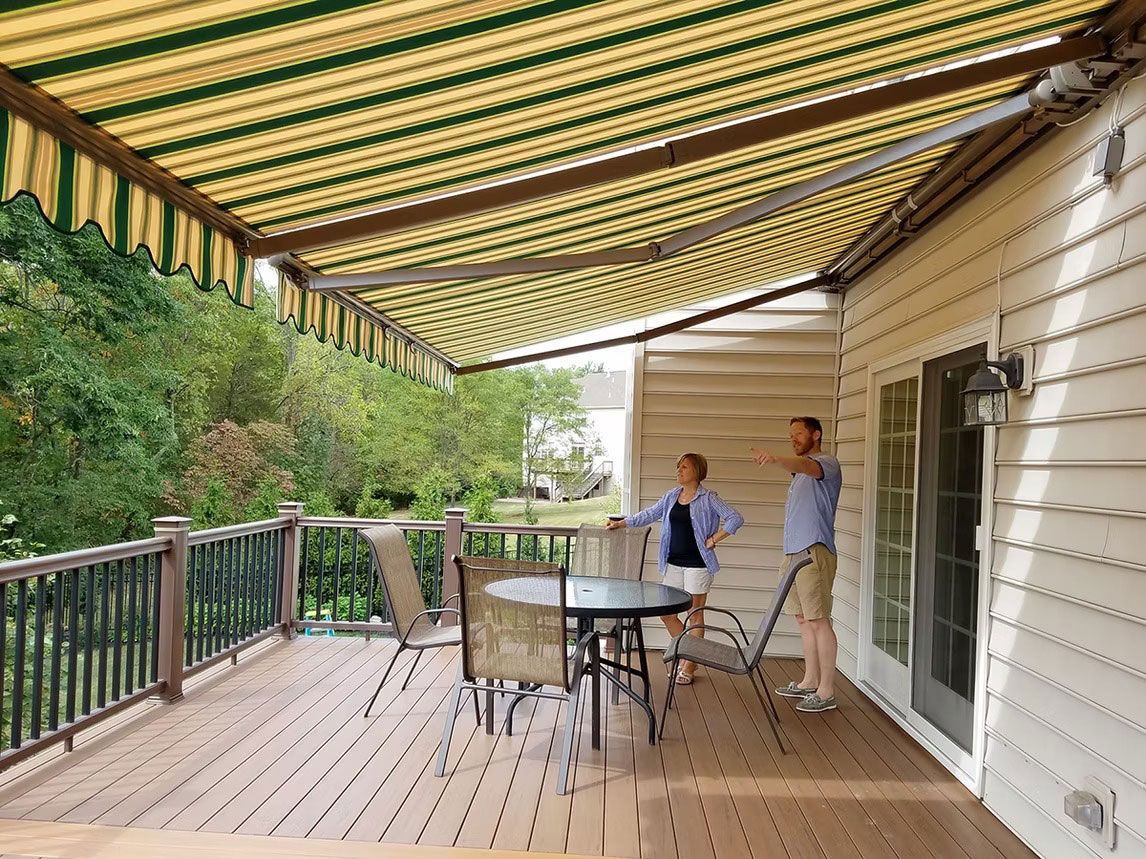 Two people stand on a deck beneath a green and yellow striped retractable awning, pointing toward a grassy yard.