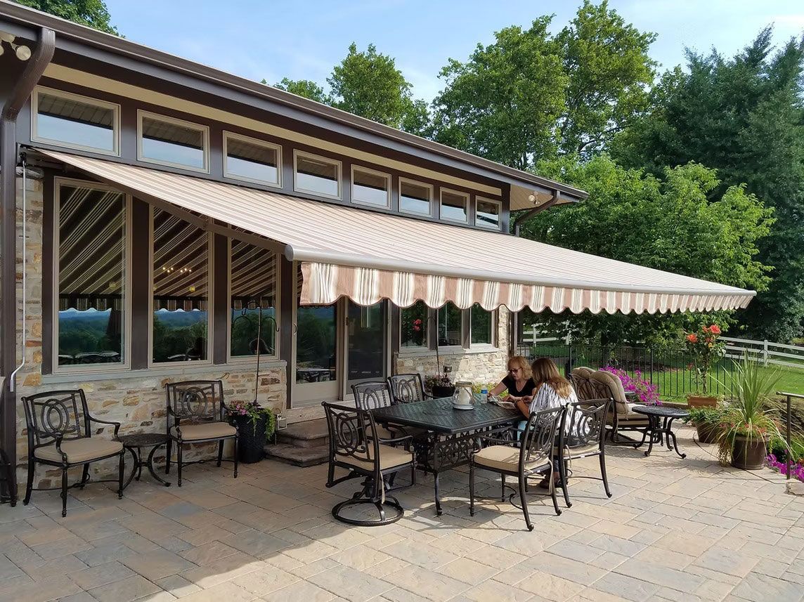 A stone patio with a striped awning extending from a house, featuring an outdoor dining set and two people seated at a table.