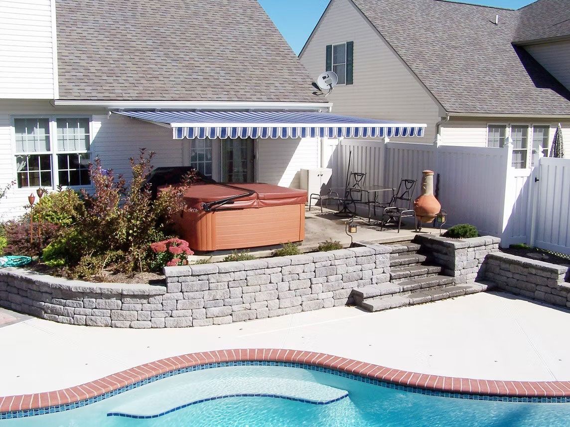 Backyard pool area with a hot tub, stone retaining wall, patio furniture, and a blue and white striped retractable awning.