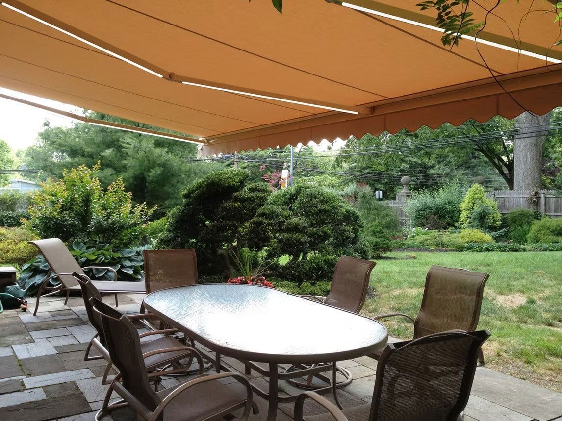 A patio table and chairs set sit under a tan retractable awning in a lush, green backyard.