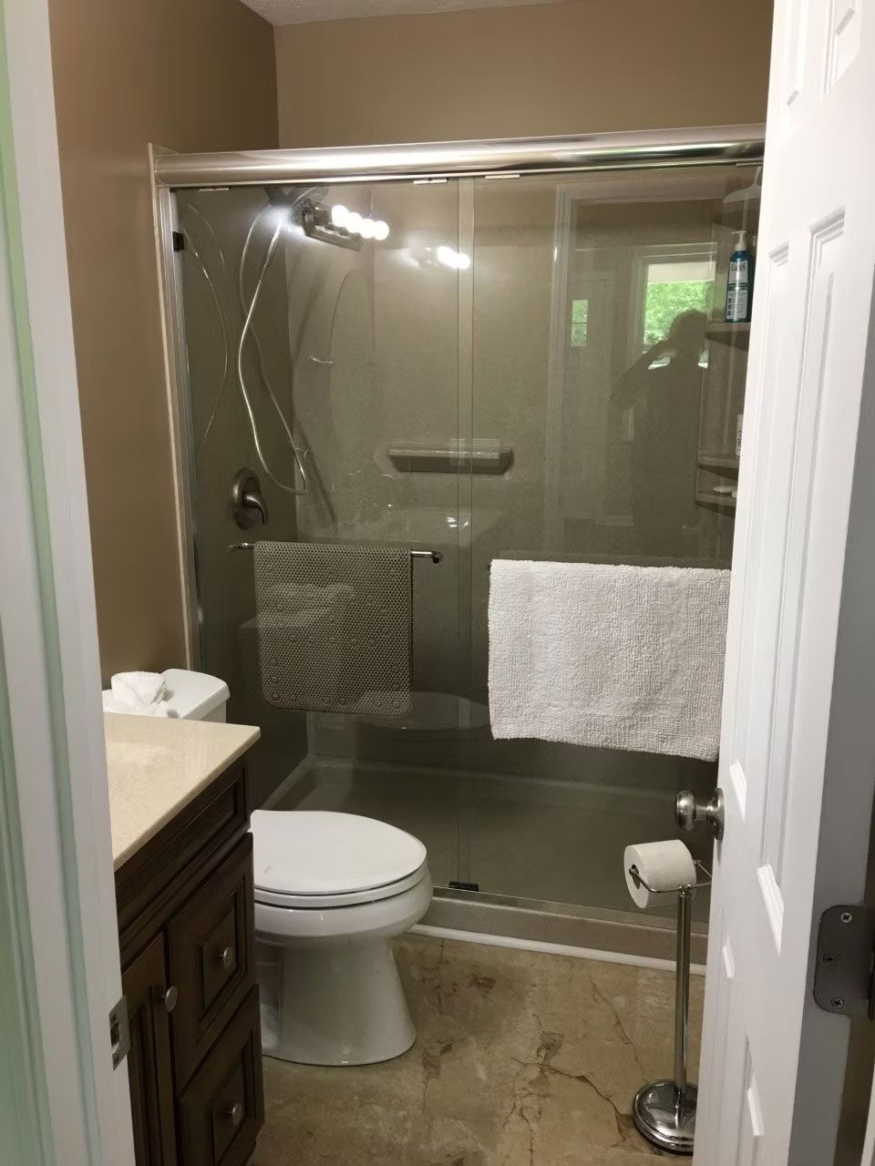 A small bathroom with a wooden vanity, white toilet, shower with glass doors, and a standalone toilet paper holder.