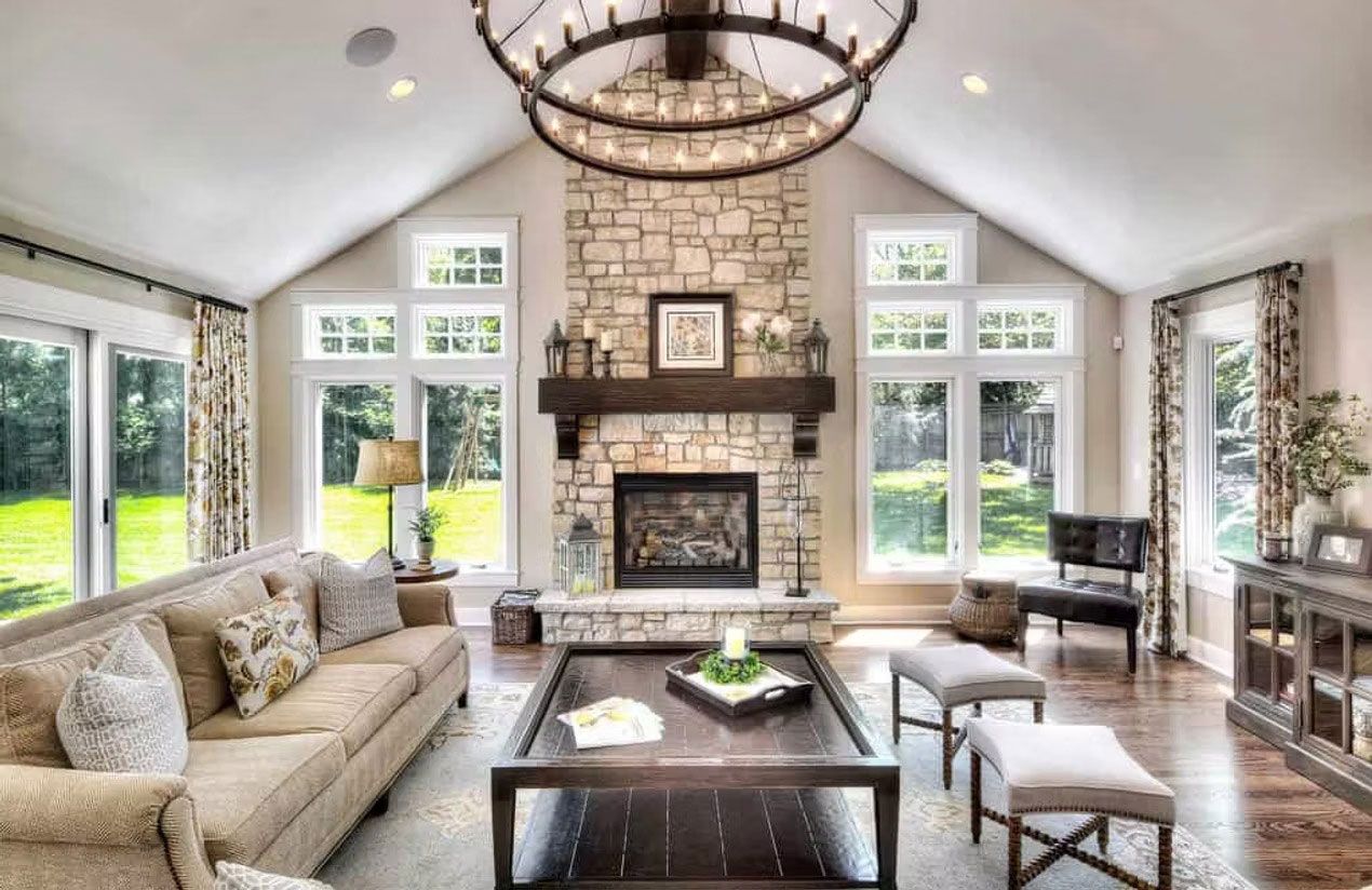 A sunlit living room featuring a stone fireplace, vaulted ceiling, large windows, a tan sofa, and a dark wooden coffee table.