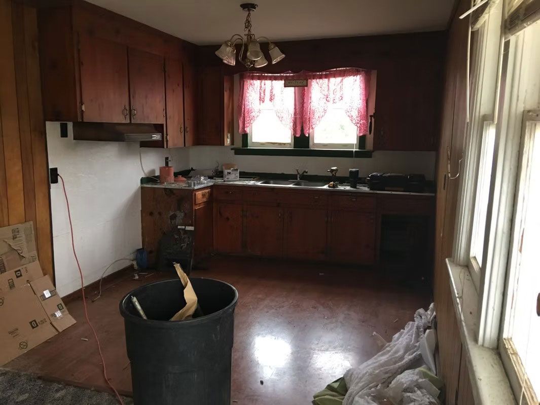 A kitchen undergoing renovation with wooden cabinets, a sink, a large black trash can, and boxes on the floor.