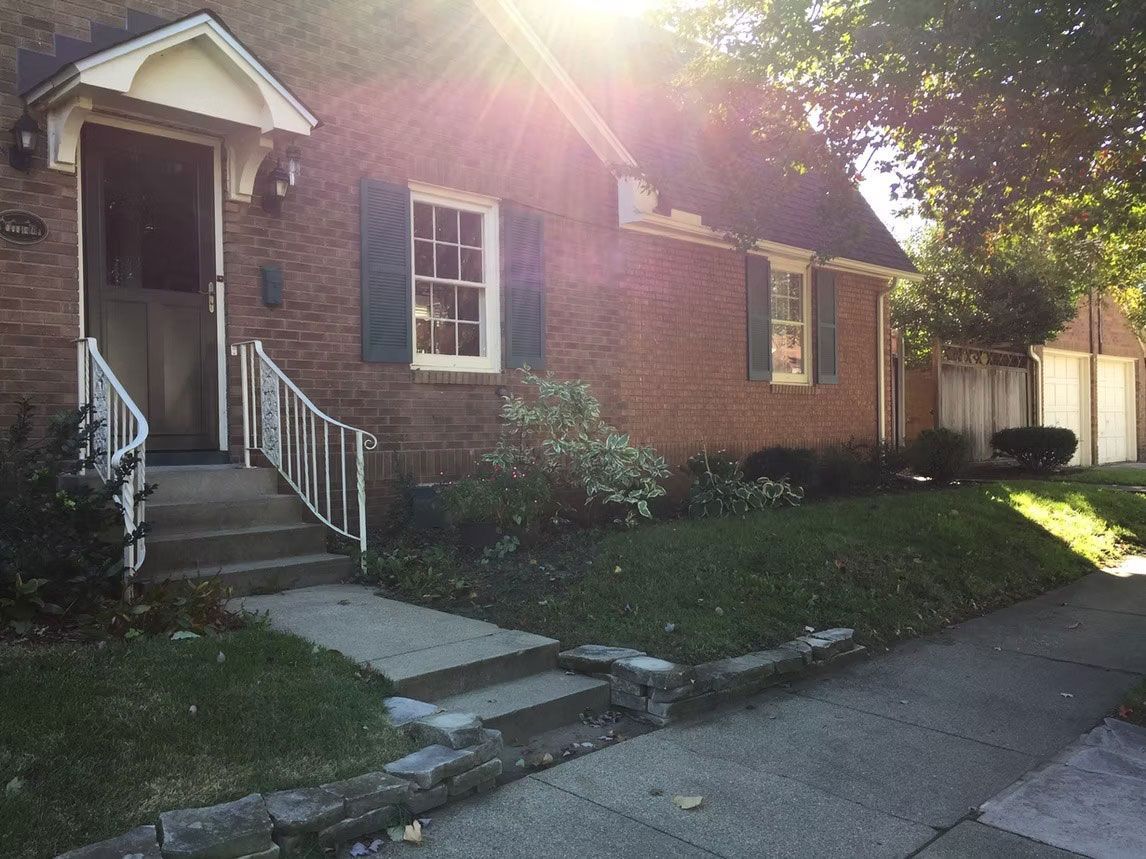 A brick house with a white front porch and stairs, green shutters, and a lawn, illuminated by sunlight.