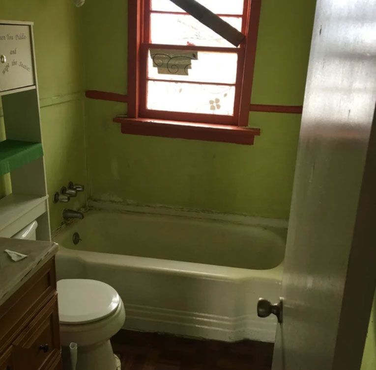A small, lime-green bathroom featuring a white porcelain bathtub, a toilet, a wooden vanity, and a window with orange trim.