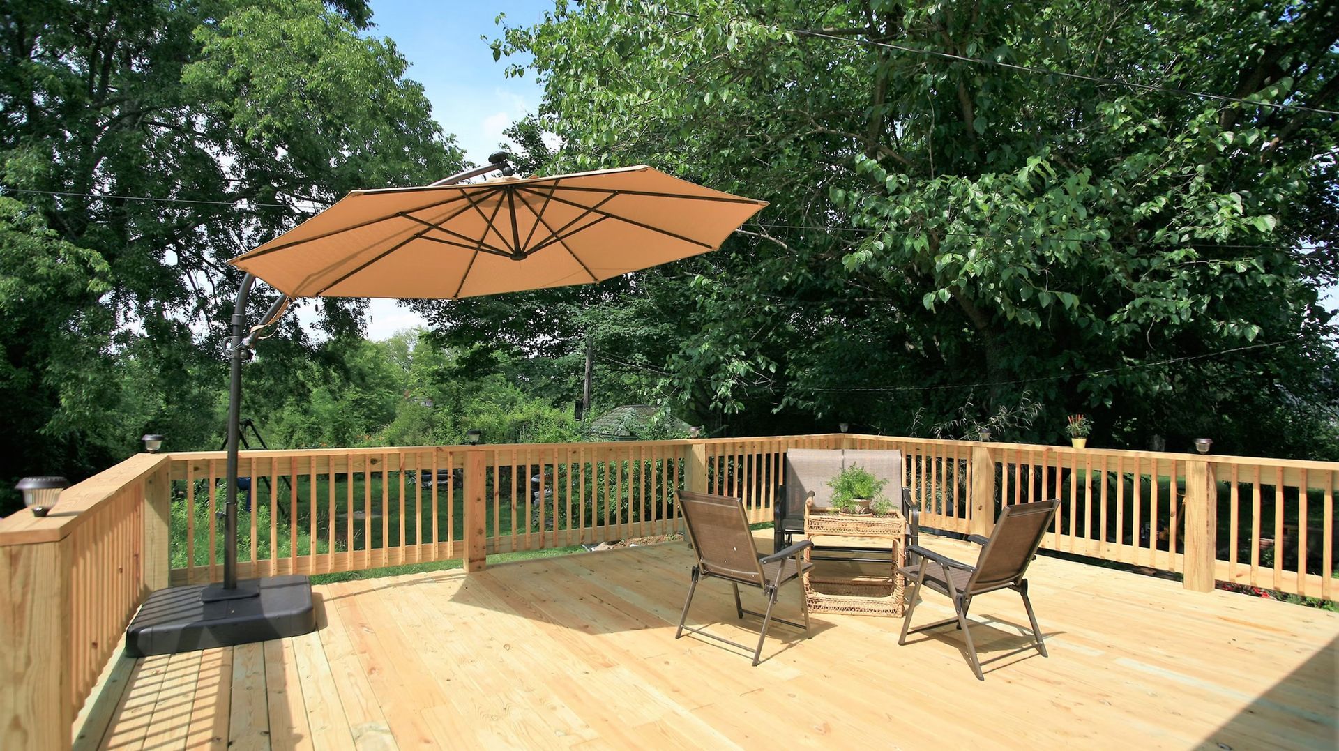A tan cantilevered patio umbrella over two chairs on a wooden deck surrounded by trees.