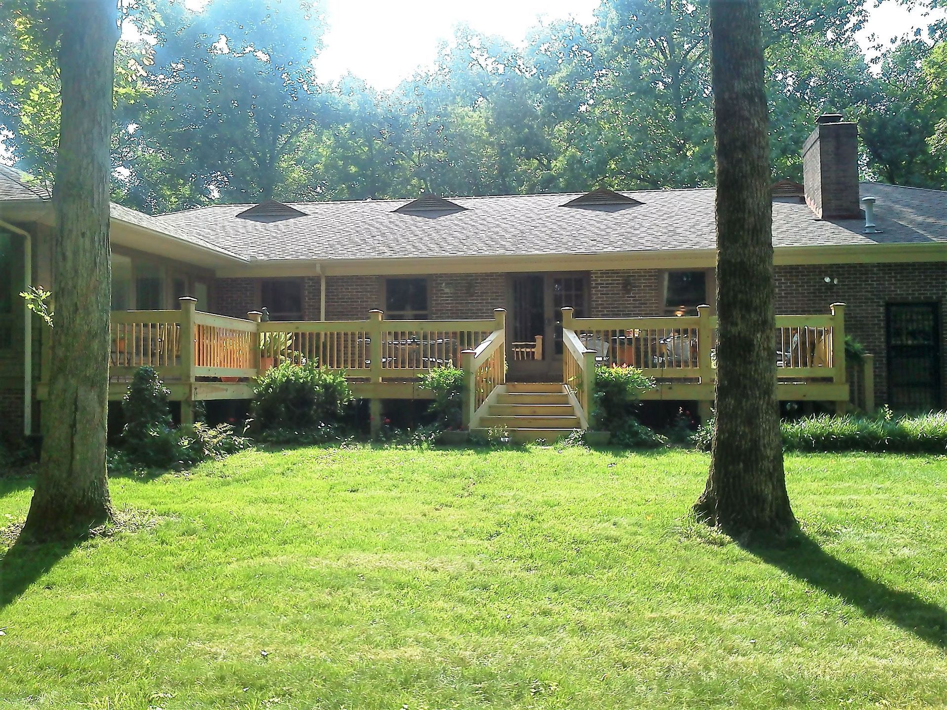 A brick house with a large wooden deck in the backyard, surrounded by green grass and trees on a sunny day.