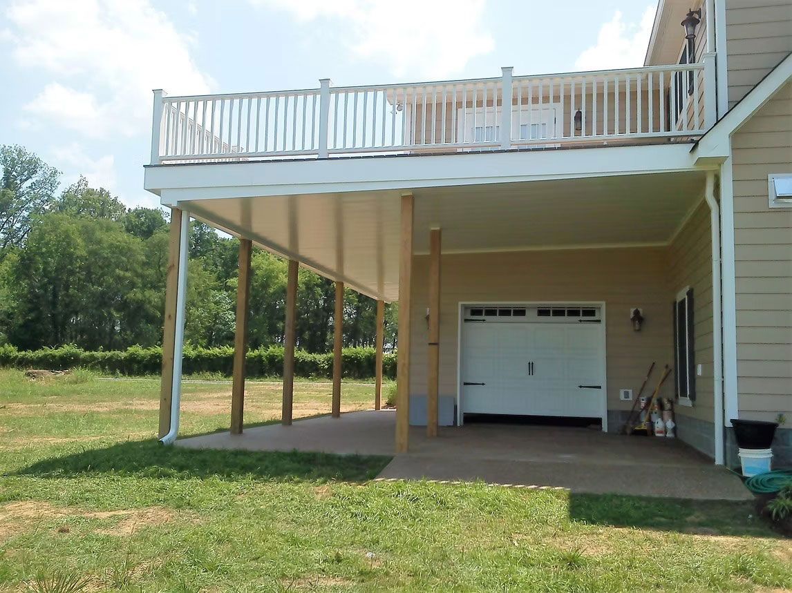 A beige house exterior featuring a large elevated deck with a white railing, supported by wooden posts above a patio.