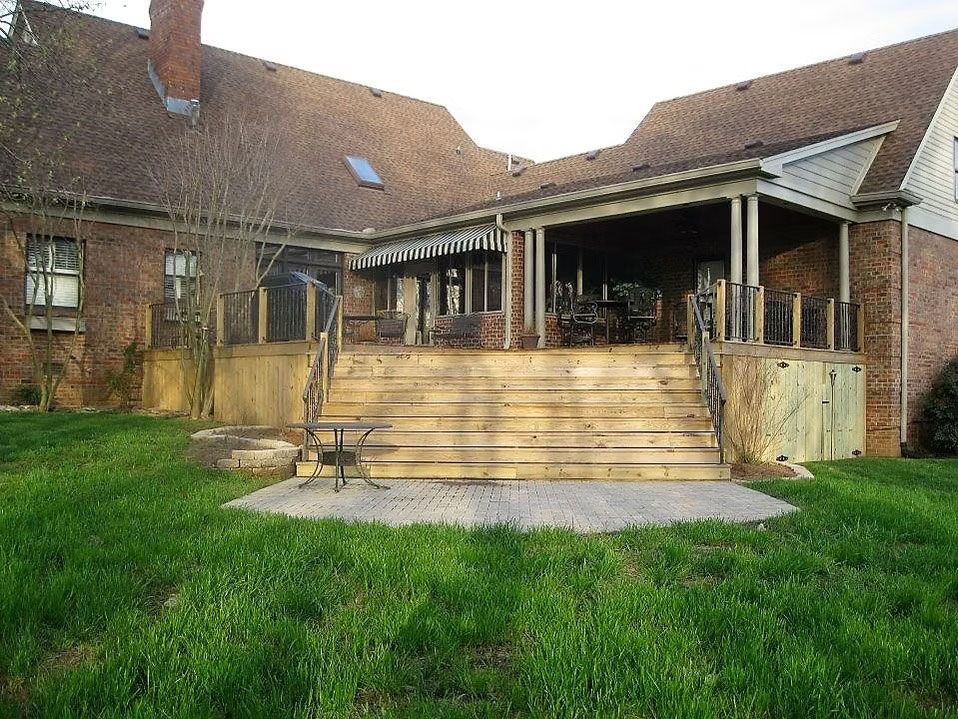 Brick house with a large wooden deck, wide staircase leading to a stone patio, and a covered porch on a grassy lawn.
