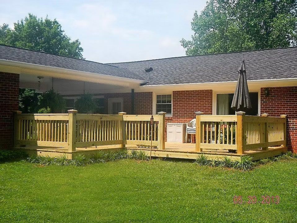 A newly built wooden deck with railings attached to a red brick house with a gray shingled roof and a backyard lawn.