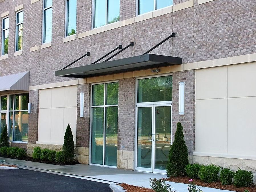 Building entrance with a modern black awning, glass double doors, and manicured landscaping in a brick storefront plaza.