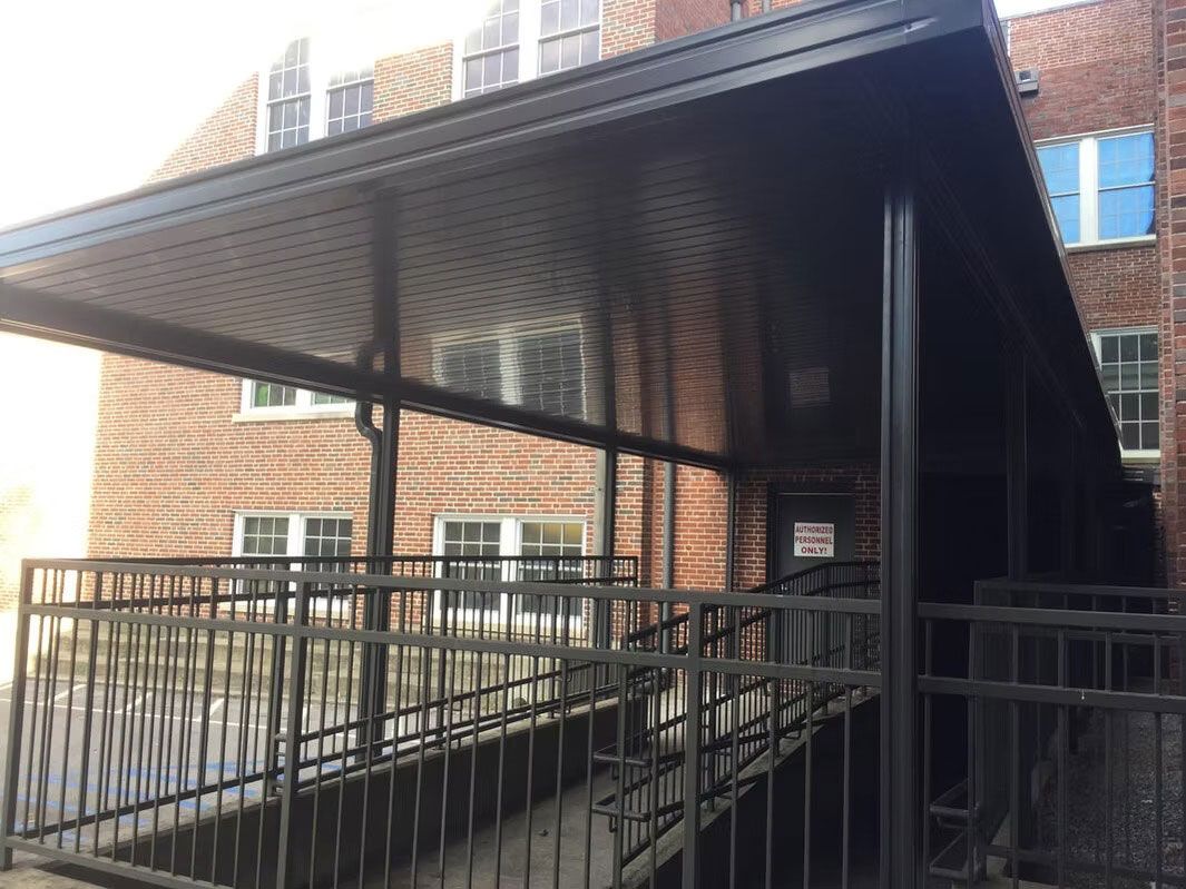 A black metal wheelchair ramp with railings leads to an entrance door under a covered walkway against a brick building.