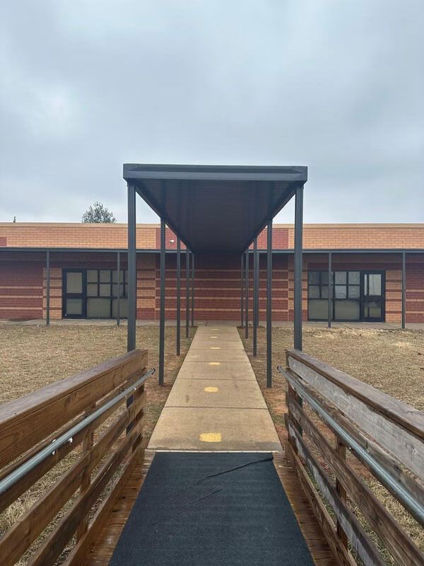 A wooden wheelchair ramp leads to a concrete walkway covered by a metal awning in front of a brick building.