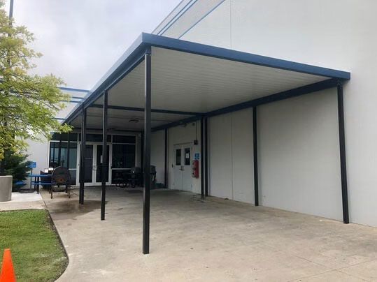 Covered walkway with a blue frame and white roof extending from a building entrance over a concrete path.