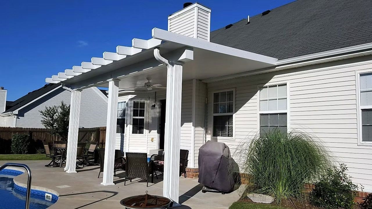 A white backyard pergola attached to a house, featuring columns, an outdoor dining area, and a covered grill near a pool.
