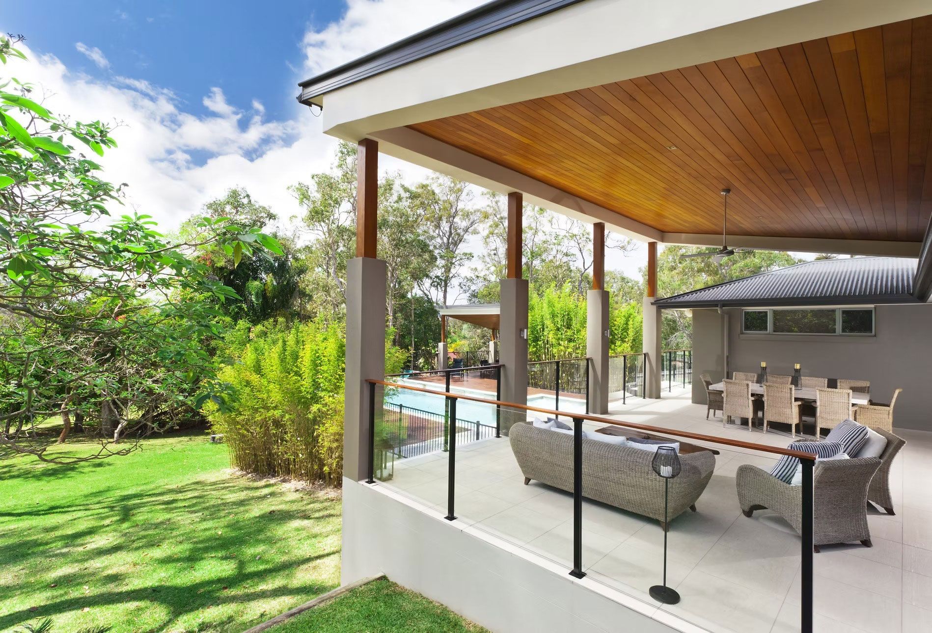 A covered outdoor patio with wicker lounge furniture and a dining set, overlooking a green garden and swimming pool.