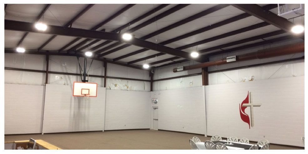 Indoor basketball court with a hoop, wooden floor, and a large red-and-black church logo on the wall