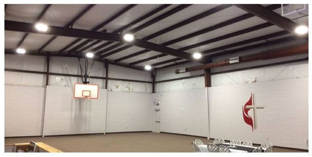 Indoor basketball court with a hoop, wooden floor, and a large red-and-black church logo on the wall