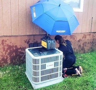 A woman is working on an air conditioner under an umbrella